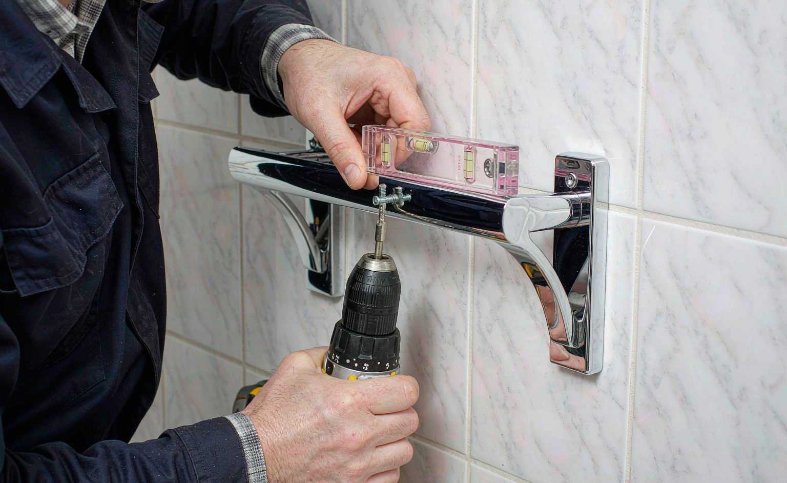 Instalador montando una correa de vidrio de ducha cromada en una pared con baldosas con una taladadora y una nivel
